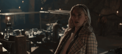 A Woman In A Plaid Coat Stands In A Dark Room Looking At The Camera GIF