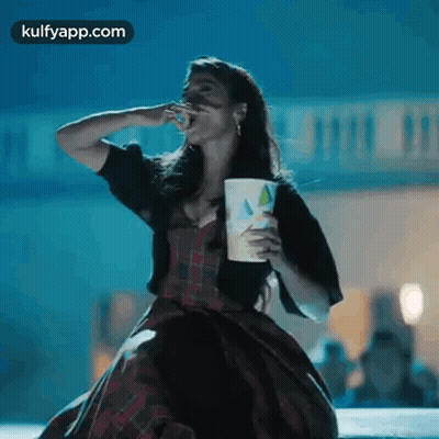 A Woman In A Plaid Dress Is Eating Popcorn And Holding A Cup . GIF