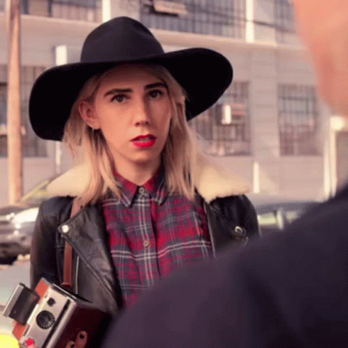 A Woman In A Plaid Shirt And A Black Hat Holds A Camera GIF
