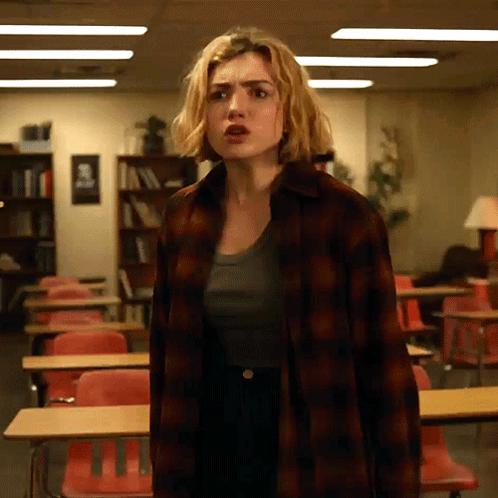 A Woman In A Plaid Shirt Stands In A Classroom With Red Chairs GIF