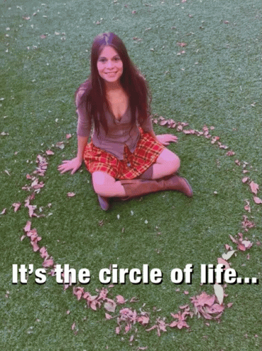 A Woman In A Plaid Skirt Sits On The Grass With Her Arms Outstretched And A Circle Of Leaves Around Her Knee GIF