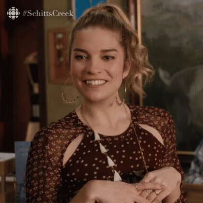 A Woman In A Polka Dot Dress Is Smiling With The #Schittscreek Logo Behind Her GIF