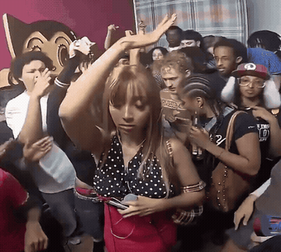 A Woman In A Polka Dot Top Is Dancing In A Crowd With A Man Wearing A Hat With The Letter O On It GIF
