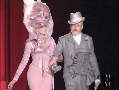A Woman In A Purple Dress And A Man In A Top Hat Are Walking Down A Runway GIF