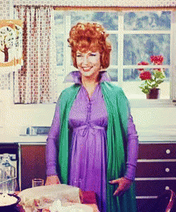 A Woman In A Purple Dress And Green Jacket Stands In Front Of A Window GIF