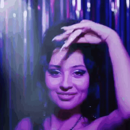 A Woman In A Purple Dress Is Smiling And Holding Her Hand To Her Forehead GIF