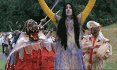 A Woman In A Purple Dress Stands In Front Of A Group Of People In Costumes GIF