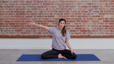 A Woman In A Purple Shirt Sits On A Blue Yoga Mat In Front Of A Brick Wall GIF