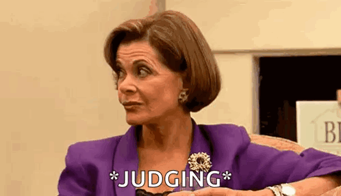 A Woman In A Purple Suit And Brooch Is Sitting In A Chair And Making A Funny Face . GIF