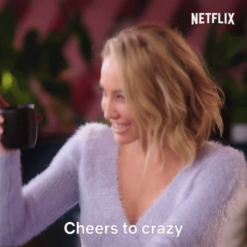 A Woman In A Purple Sweater Is Holding A Cup Of Coffee And Says " Cheers To Crazy " GIF