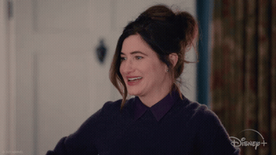 A Woman In A Purple Sweater Is Smiling In Front Of A Sign That Says Disney + GIF