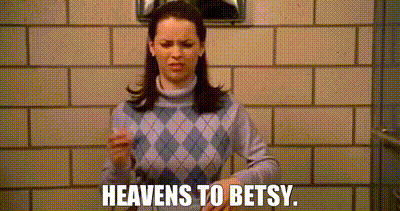 A Woman In A Purple Sweater Is Standing In Front Of A Brick Wall And Saying `` Heavens To Betsy '' . GIF