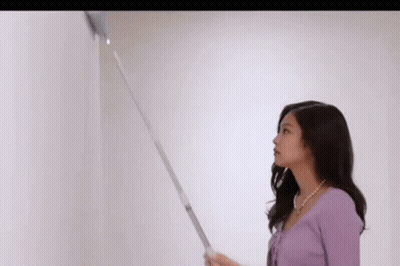 A Woman In A Purple Sweater Is Standing Next To A White Wall And Holding A Vacuum Cleaner . GIF
