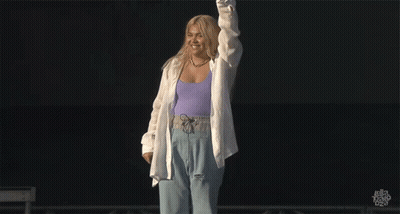 A Woman In A Purple Tank Top And White Shirt Stands On A Stage GIF
