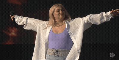 A Woman In A Purple Top And White Shirt Is Holding A Microphone With Her Arms Outstretched GIF