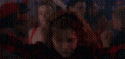 A Woman In A Red And Black Plaid Shirt Is Dancing In Front Of A Crowd . GIF