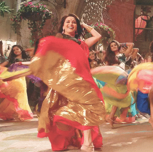 A Woman In A Red And Gold Dress Is Dancing With Other Women In Colorful Dresses GIF
