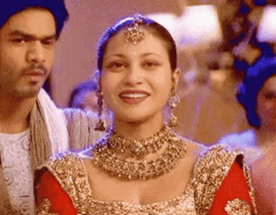 A Woman In A Red And Gold Dress Is Smiling While Standing Next To A Man . GIF