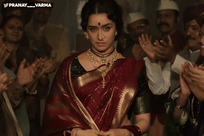 A Woman In A Red And Gold Saree Stands In Front Of A Crowd Of People With The Words Pranay Varma Above Her GIF