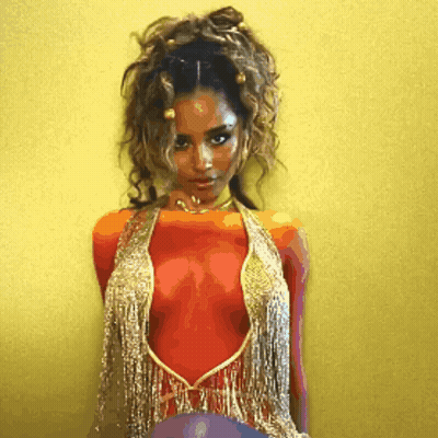 A Woman In A Red And Gold Top With Fringe Is Standing In Front Of A Yellow Wall GIF