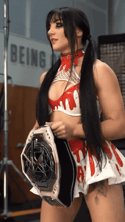 A Woman In A Red And White Outfit Is Holding A Wrestling Belt In Front Of A Sign That Says Being GIF