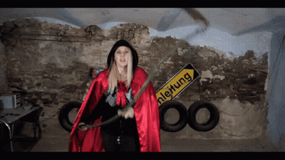 A Woman In A Red Cape Stands In Front Of A Yellow Sign That Says Leitung GIF