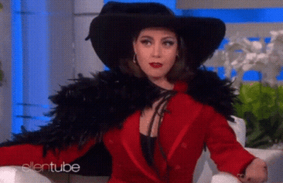 A Woman In A Red Coat And Black Hat Is Sitting On A Chair . GIF