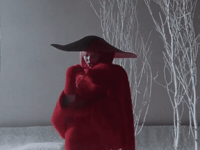 A Woman In A Red Coat And Black Hat Stands In Front Of Snowy Trees GIF