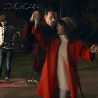 A Woman In A Red Coat Is Dancing With A Man Playing An Accordion And The Words Love Again Are Above Them GIF