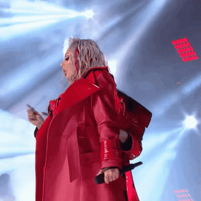 A Woman In A Red Coat Is Singing Into A Microphone On A Stage GIF