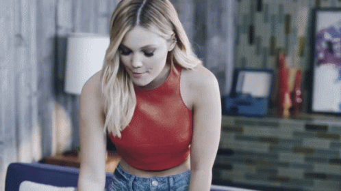 A Woman In A Red Crop Top And Blue Jeans Is Sitting On A Couch GIF