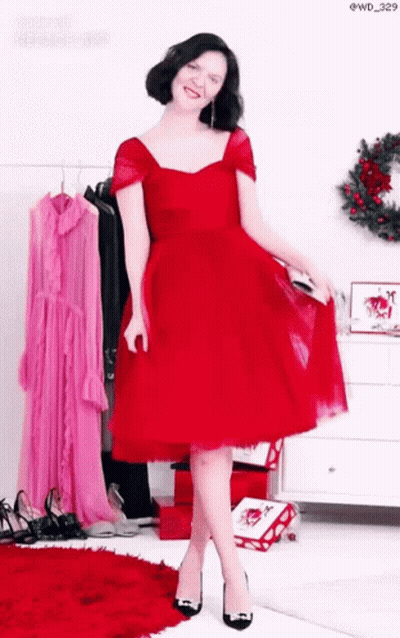 A Woman In A Red Dress And Black Heels Is Standing In Front Of A Wreath . GIF
