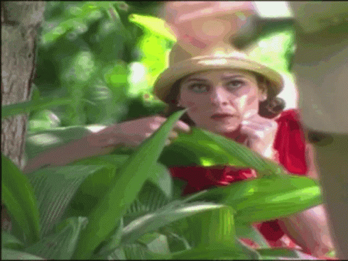A Woman In A Red Dress And Straw Hat Looks Out From Behind Some Green Plants GIF