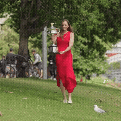 A Woman In A Red Dress Is Carrying A Trophy In A Park GIF