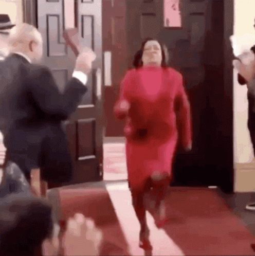 A Woman In A Red Dress Is Running On A Red Carpet While A Man In A Suit Stands Behind Her . GIF