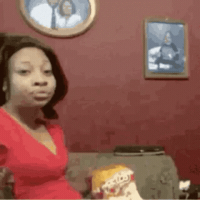 A Woman In A Red Dress Is Sitting On A Couch With A Picture Of A Man On The Wall Behind Her . GIF