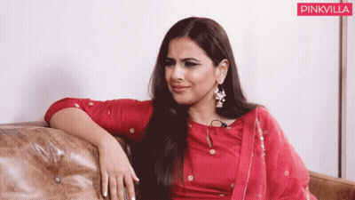 A Woman In A Red Dress Is Sitting On A Couch With A Pinkvilla Logo In The Corner GIF