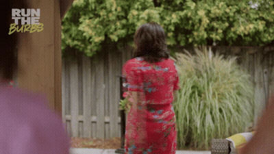 A Woman In A Red Dress Is Standing In Front Of A Fence With The Words Run The Burbs On The Bottom GIF