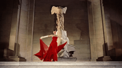 A Woman In A Red Dress Is Standing In Front Of A Large Statue GIF