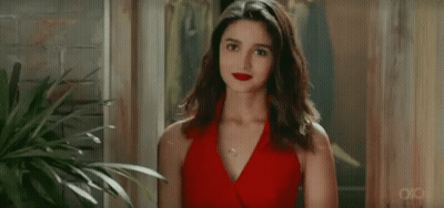 A Woman In A Red Dress Is Standing In Front Of A Plant And Looking At The Camera . GIF