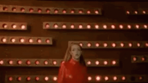 A Woman In A Red Dress Is Standing In Front Of A Wall Of Lights And A Sign That Says Oops . GIF