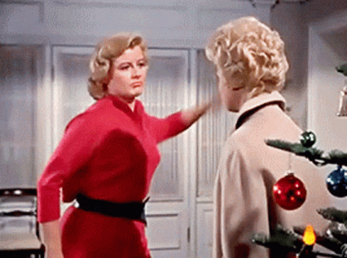 A Woman In A Red Dress Is Standing Next To A Man In A Tan Coat Near A Christmas Tree . GIF