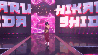 A Woman In A Red Dress Is Standing On A Stage In Front Of A Large Screen That Says ' Nikki Shida ' GIF