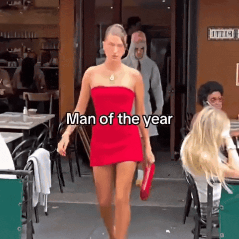 A Woman In A Red Dress Is Walking In Front Of A Restaurant With The Words Man Of The Year On The Bottom GIF