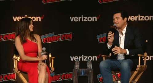 A Woman In A Red Dress Sits Next To A Man Holding A Microphone At A Comic Con GIF