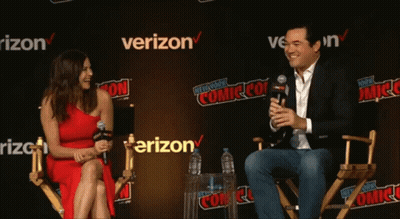 A Woman In A Red Dress Sits Next To A Man In A Suit In Front Of A Verizon Sign GIF