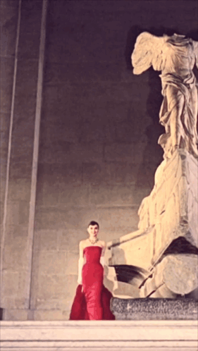 A Woman In A Red Dress Stands Next To A Statue GIF