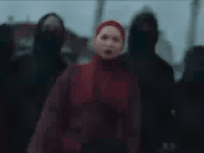 A Woman In A Red Hijab Is Standing In Front Of A Group Of People In Black Hoods . GIF