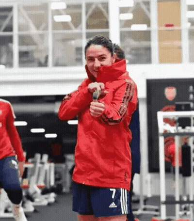 A Woman In A Red Jacket And Blue Shorts Is Standing In A Gym Holding A Microphone . GIF