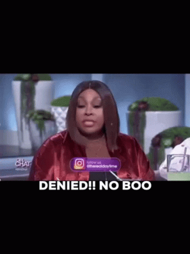 A Woman In A Red Jacket Is Sitting At A Table With A Sign That Says `` Denied ! No Boo '' . GIF